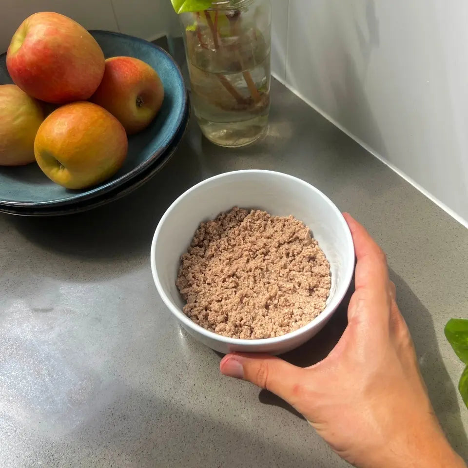 Hand holding a bowl of Louis fresh cat food on a kitchen bench beside apples and a vase.